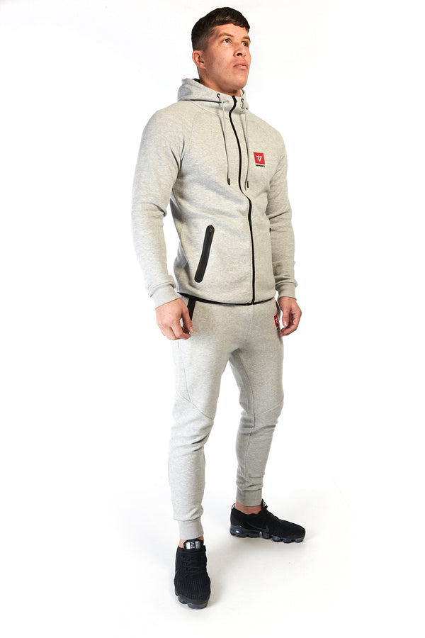 Man wearing grey, slim fit tracksuit pants with red logo on the left thigh